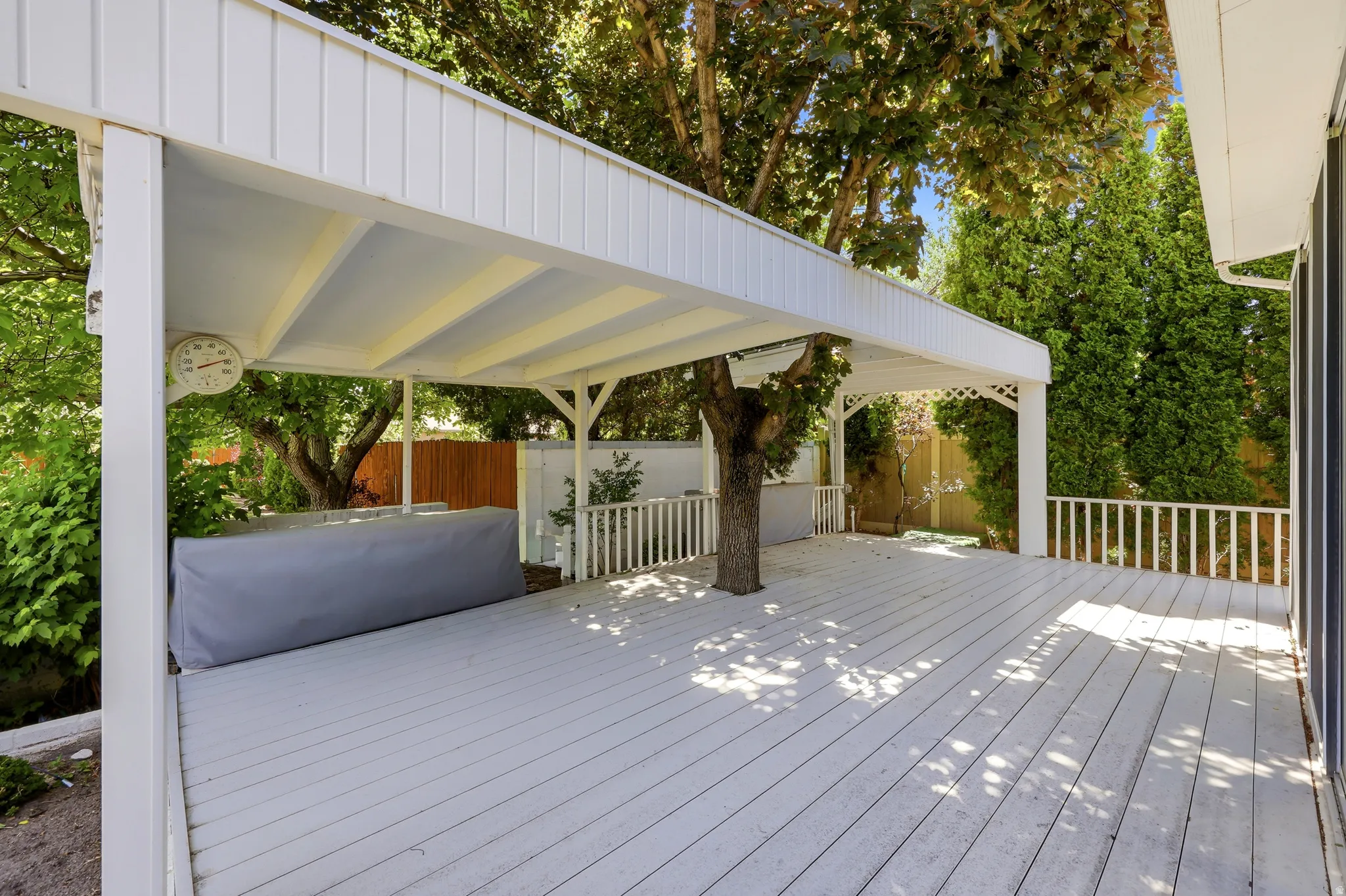 Expansive outdoor deck featuring a white painted surface, a partial overhead patio cover, white railings, mature trees, and privacy fencing