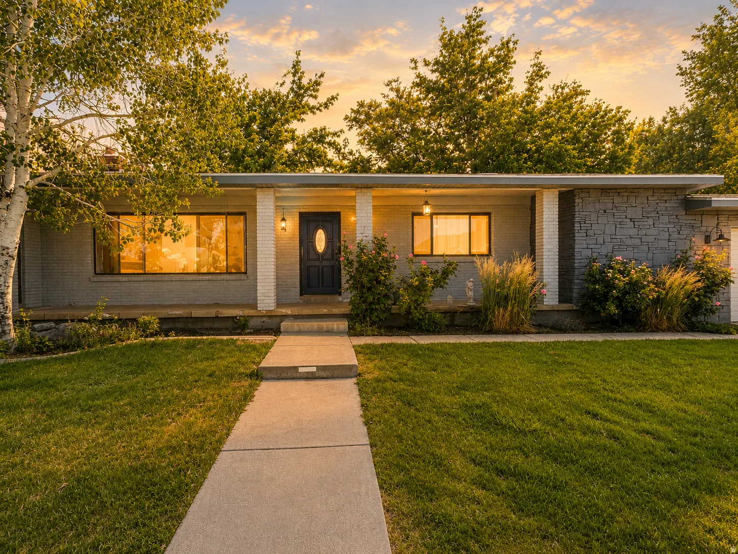 Mid-century modern exterior featuring a gray brick facade, stone accent walls, a covered entry with white support columns, an oval-paneled front door, and a concrete walkway