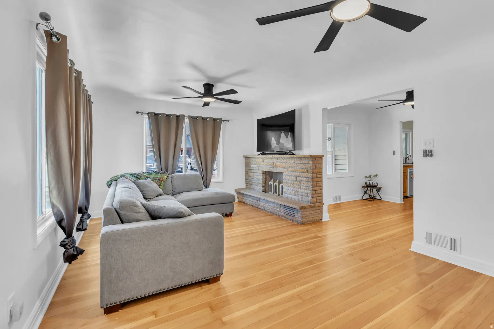 Spacious living area featuring wood-finish flooring, a stone-front fireplace, multiple windows, and contemporary ceiling fans