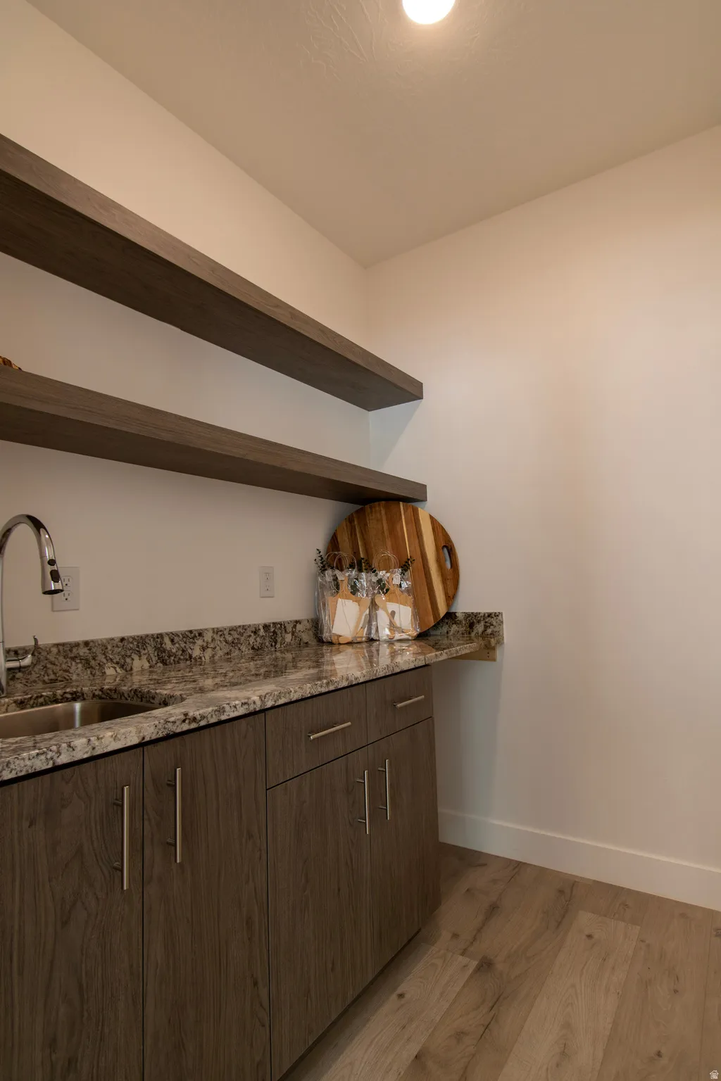 Utility area featuring a stainless steel sink, patterned countertop, wood-finish cabinetry with brushed nickel hardware, and two floating shelves