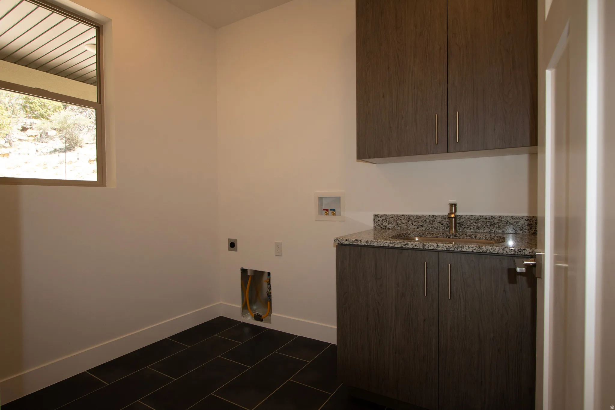 Utility room featuring dark wood-finish cabinetry, granite countertop with undermount sink, and dark tile flooring