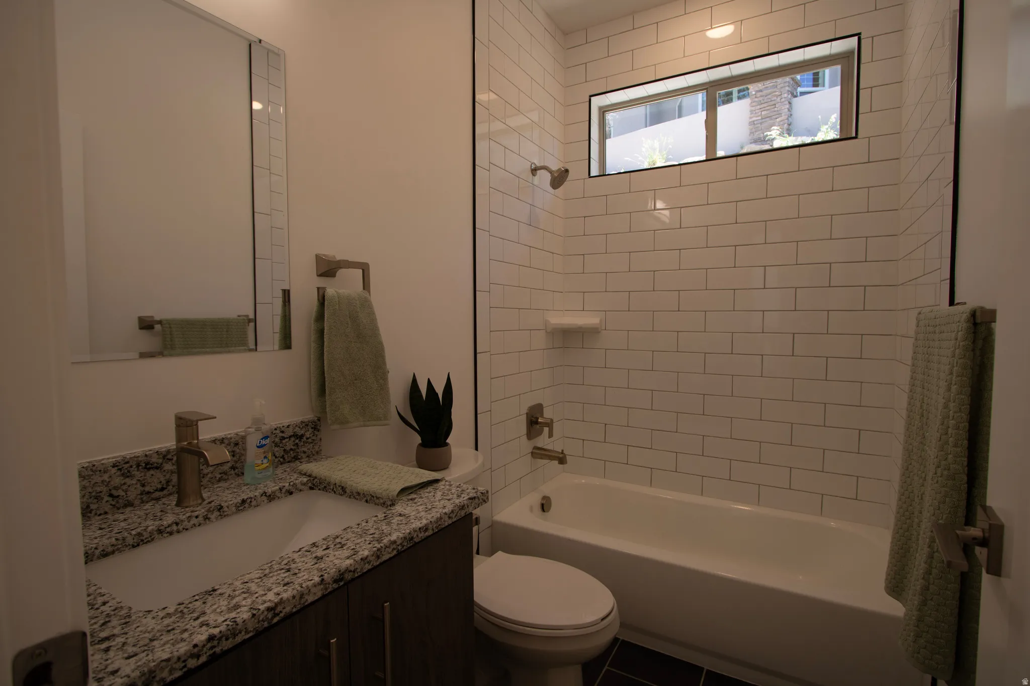 Bathroom featuring a white subway tile surround, an undermount sink with a granite-look countertop, a dark wood-finish vanity, a built-in bathtub, and a horizontal window
