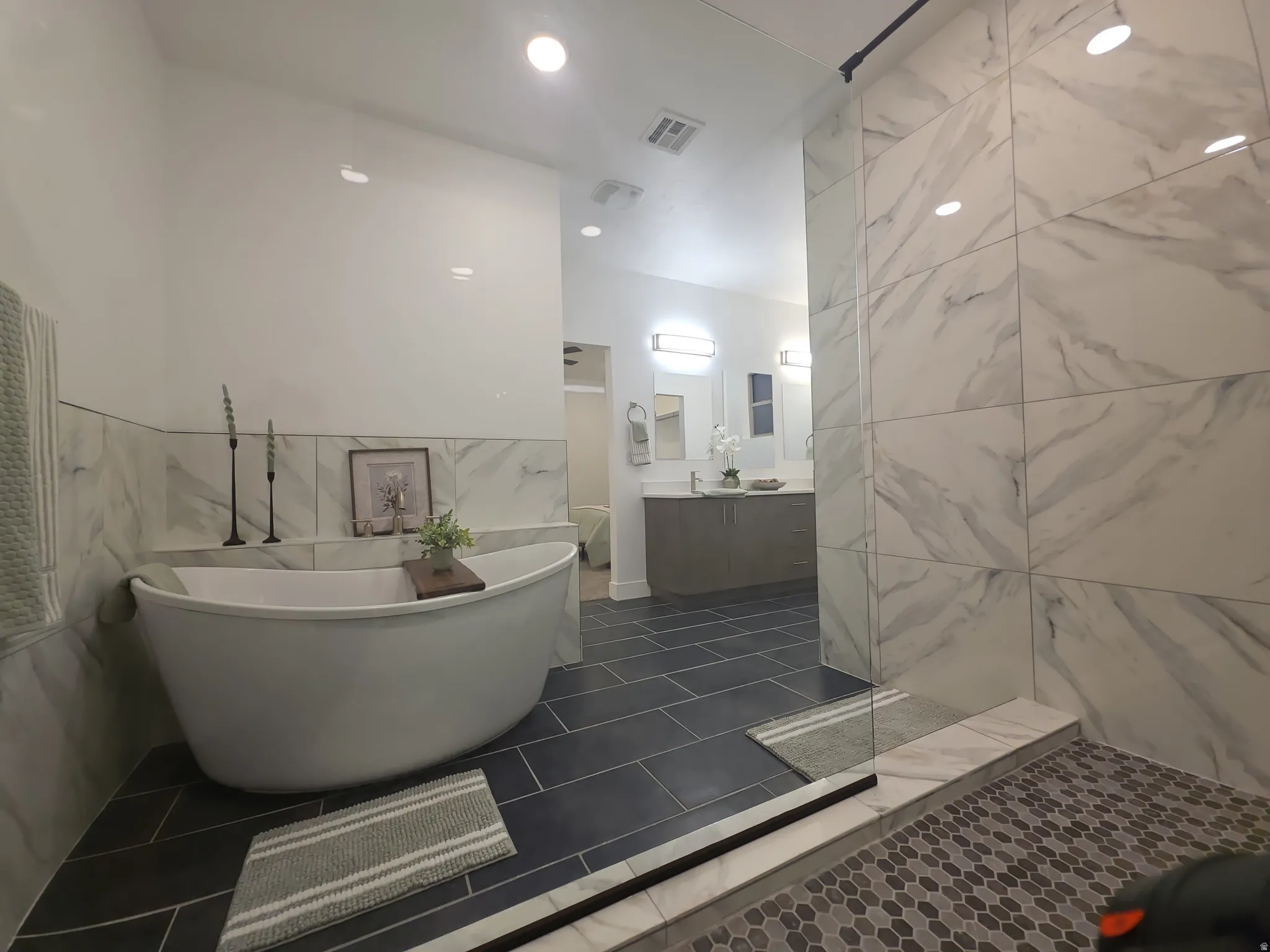 Spacious bathroom featuring a freestanding soaking tub, large format tile shower, dual vanity with undermount sinks, and contrasting floor tiles