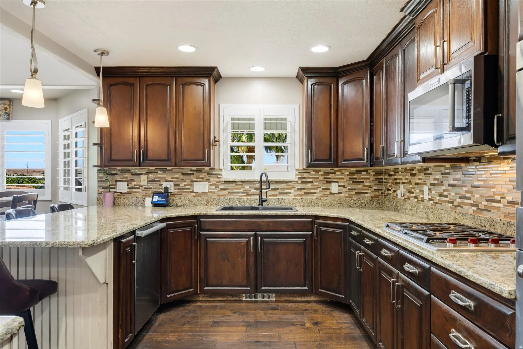 Kitchen featuring wood-finish flooring, dark wood cabinetry, light granite countertops, a tiled backsplash, and stainless steel appliances including a microwave and gas range