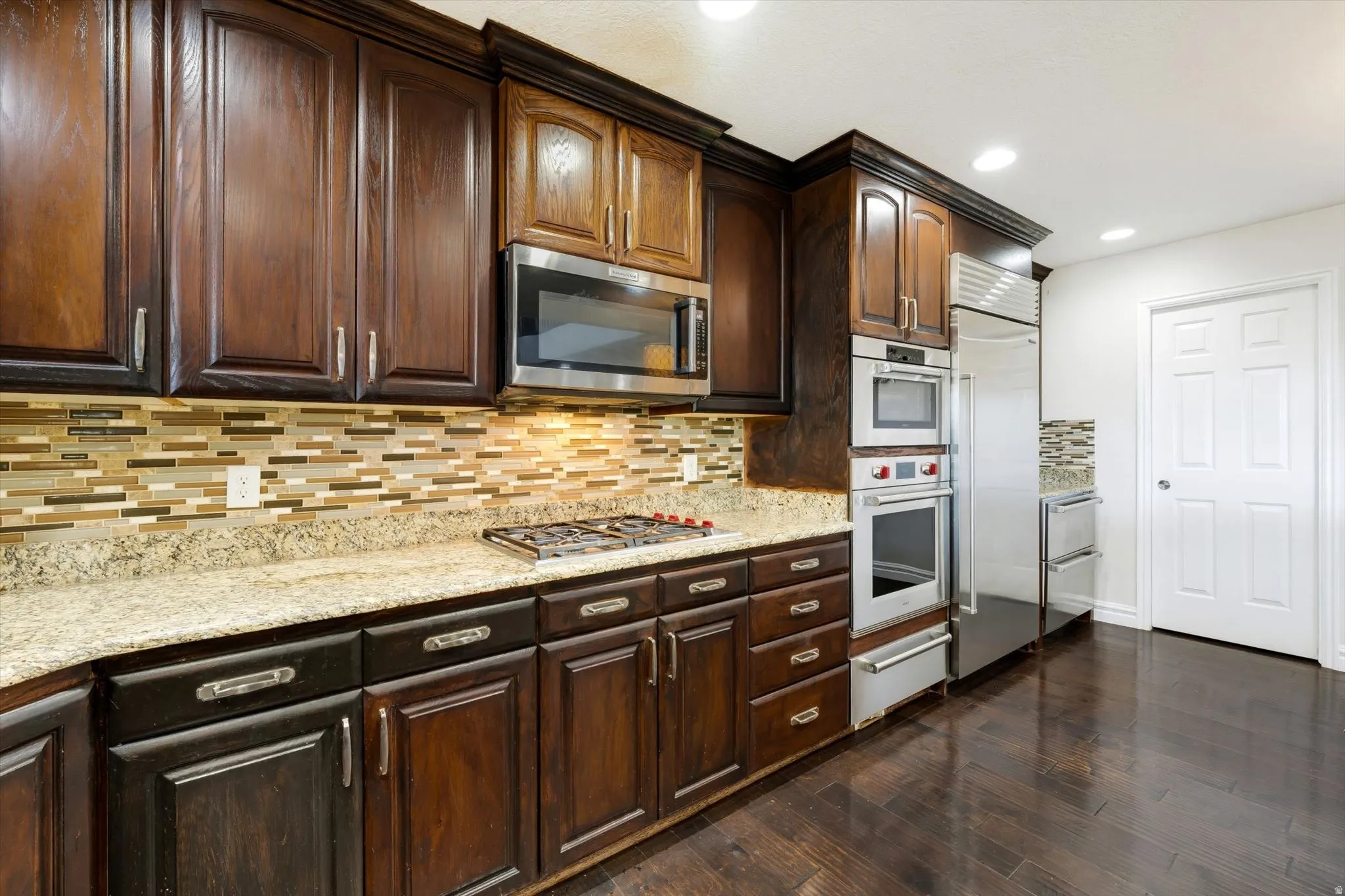 Gourmet kitchen featuring rich wood cabinetry, granite countertops, a mosaic tile backsplash, stainless steel appliances, and dark wood-finish flooring