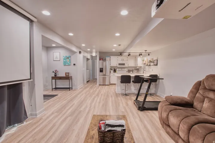 Recessed lighting illuminates wood-finish flooring throughout this open-concept space featuring a built-in projection screen, a kitchen with an island and integrated stainless steel appliances, and a hallway entrance