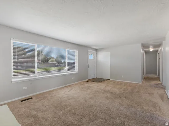 Spacious room with plush carpeting, neutral grey wall paint, and a large front window with blinds