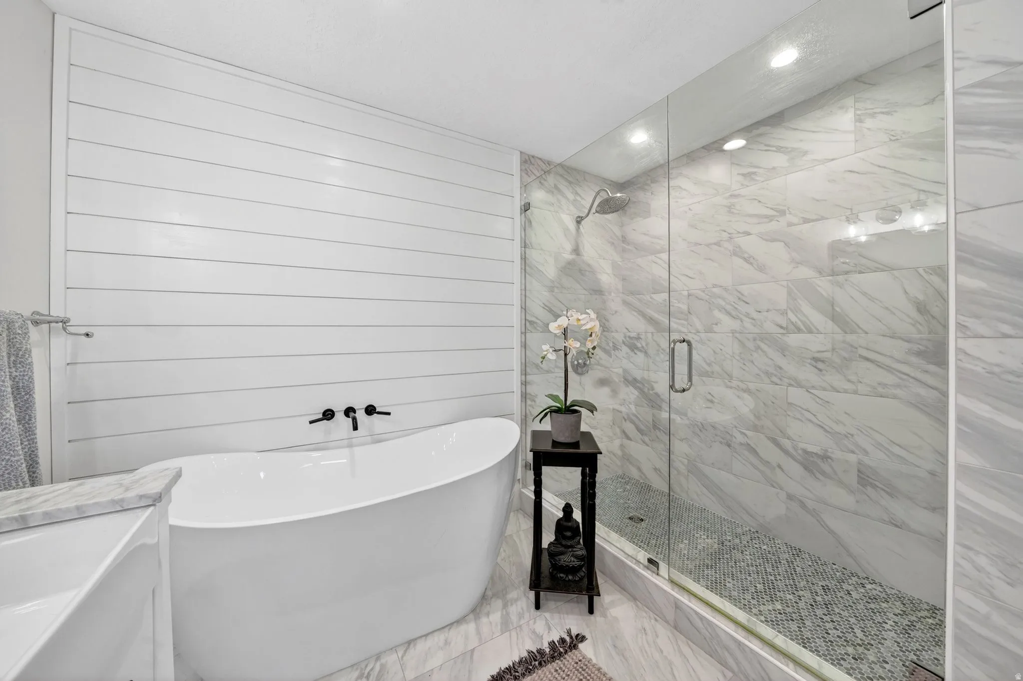 Contemporary bathroom featuring a freestanding soaking tub with wall-mounted matte black fixtures, a shiplap accent wall, and a spacious glass-enclosed shower with large format marble-look tile walls and a mosaic pebble floor