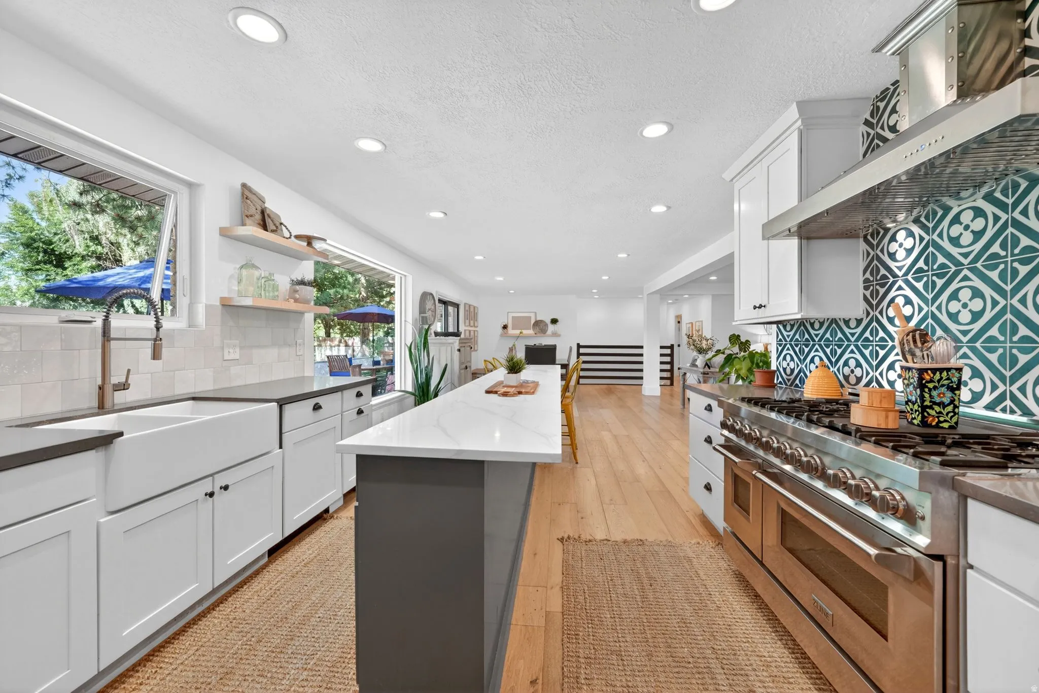 Contemporary kitchen featuring an oversized island, stainless steel range with custom tile backsplash, farmhouse sink, recessed lighting, and wood-finish flooring