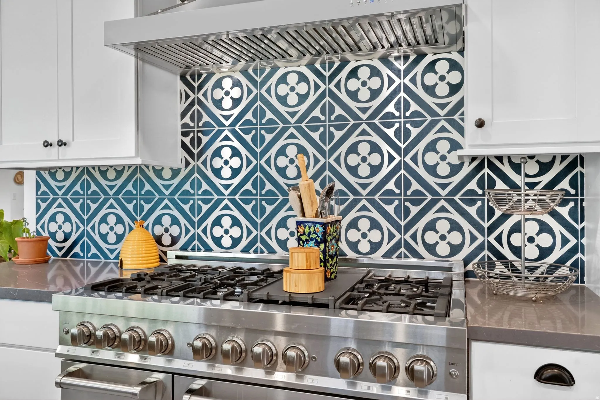 Stainless steel range with multiple burners, patterned tile backsplash, white upper cabinetry, and dark countertops