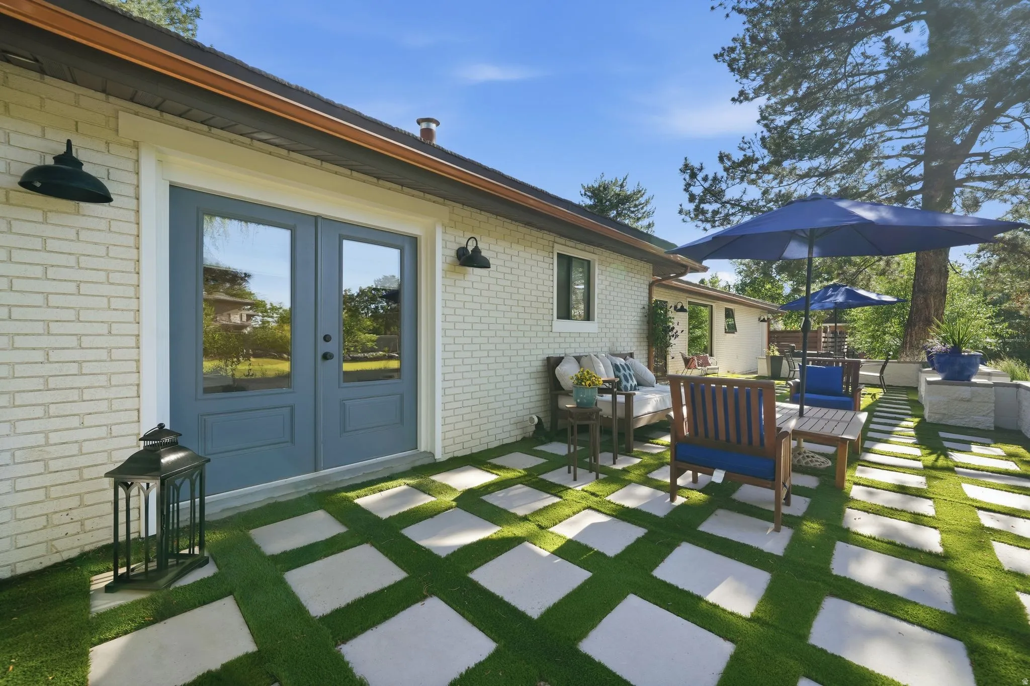 Rear patio with light-colored brick exterior, featuring blue double doors, a geometric paver and artificial turf pattern, and a built-in planter