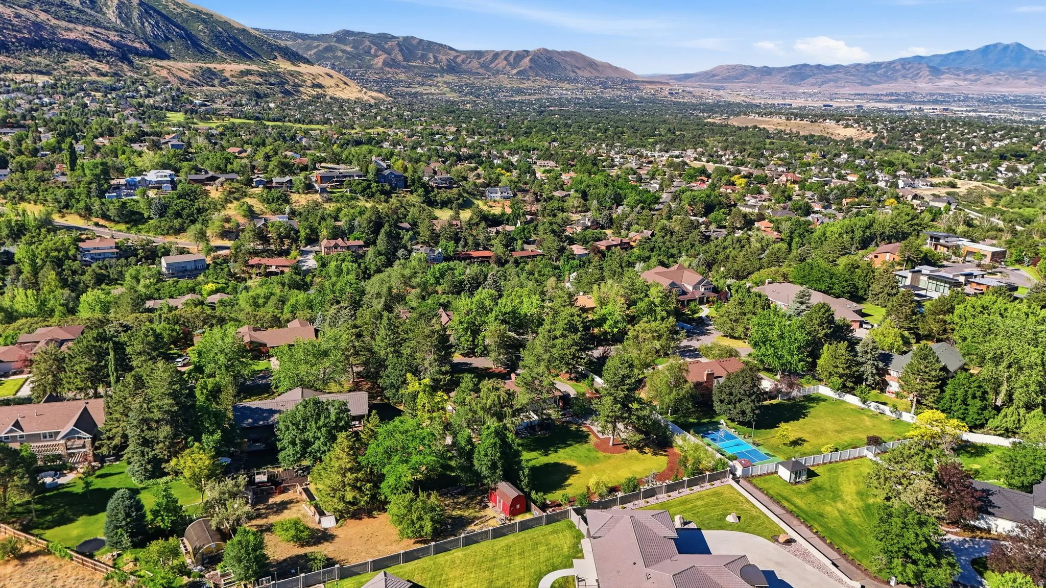 Expansive property with a tennis court, manicured lawn, and mature trees, surrounded by residential properties and distant mountains