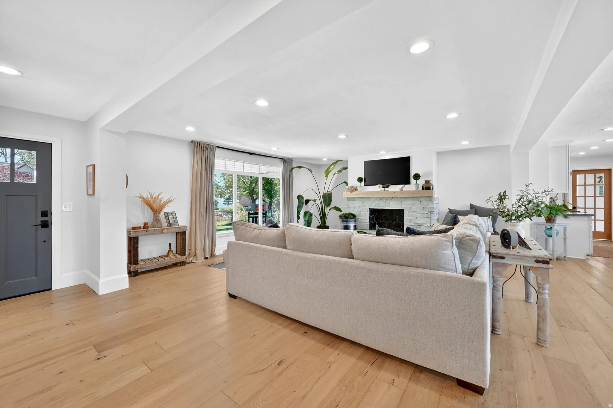 Spacious living area featuring light wood-finish flooring, recessed lighting, a stone-clad fireplace with a wood mantel, and large windows with drapery