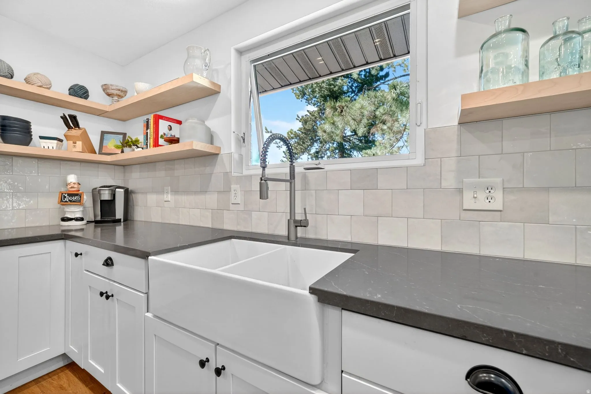 Kitchen featuring a dual-basin farmhouse sink with a pull-down faucet, dark countertops, white cabinetry, and a light-toned tile backsplash