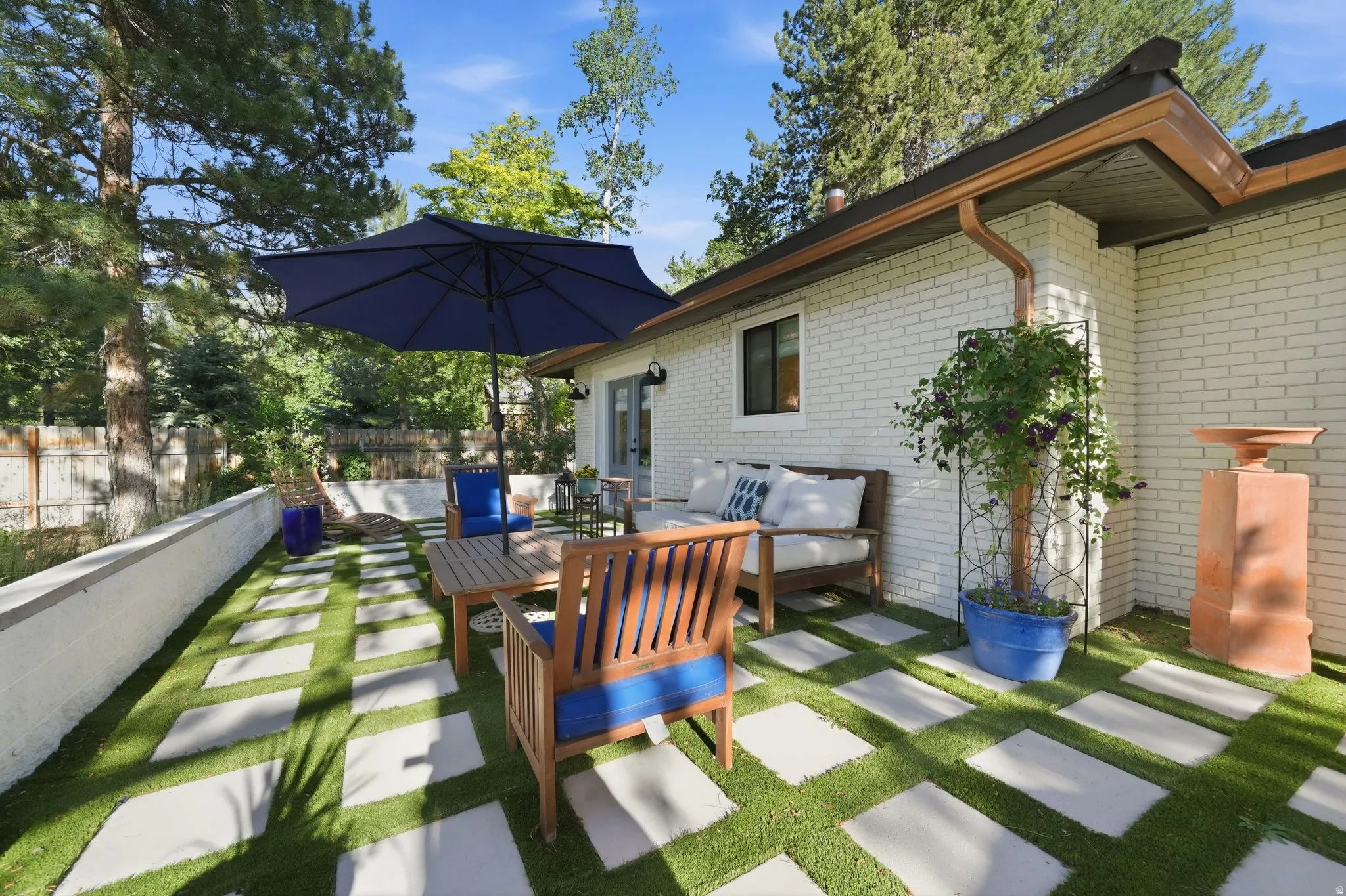 Outdoor patio featuring rectangular pavers set in artificial turf, a white brick exterior, a white retaining wall, and mature perimeter trees