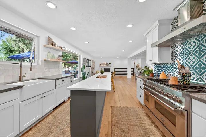 Contemporary kitchen featuring an oversized island, stainless steel range with custom tile backsplash, farmhouse sink, recessed lighting, and wood-finish flooring