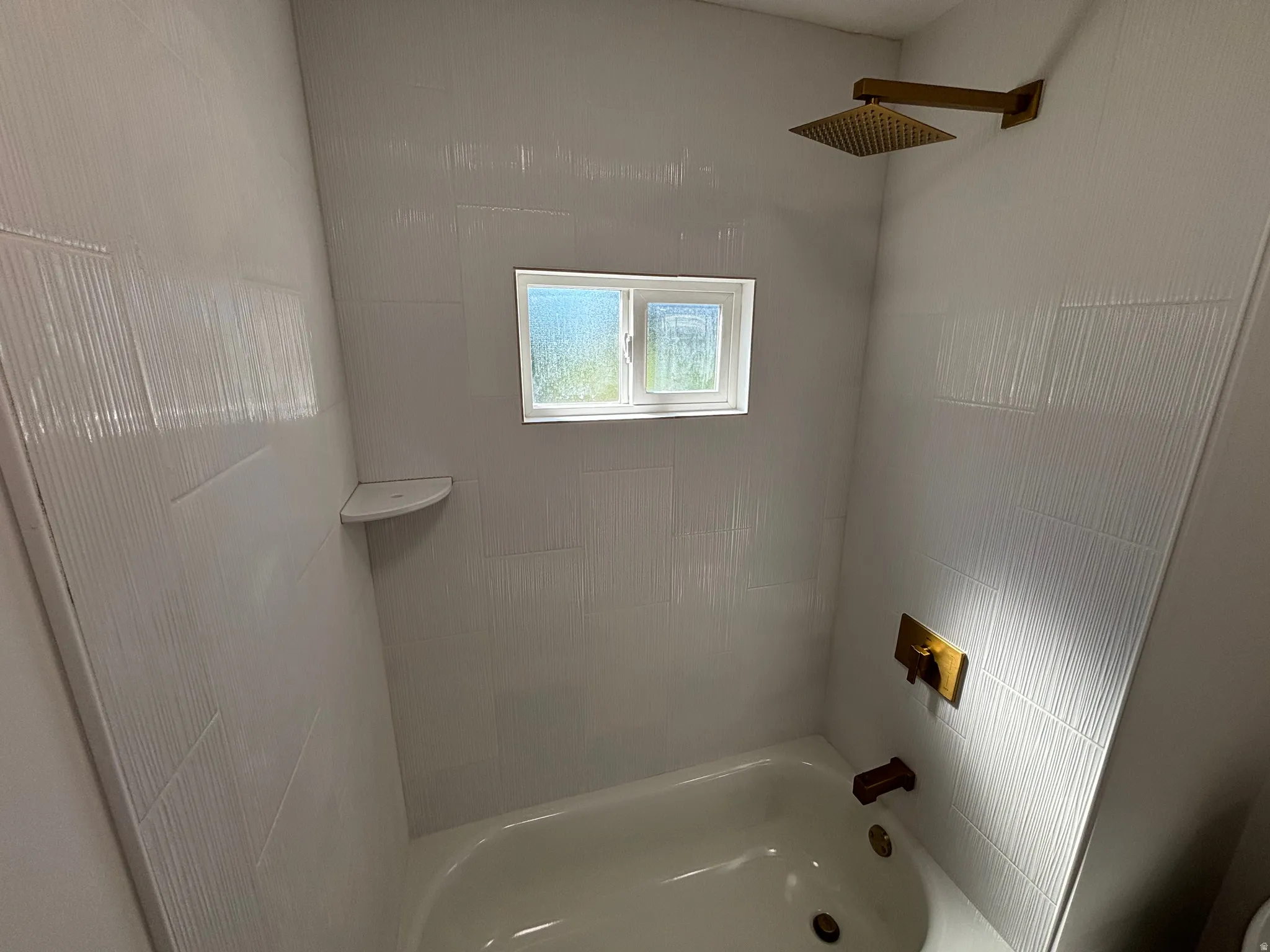 Bathtub and shower combination featuring white textured tile surround, a built-in corner shelf, and a window with privacy glass