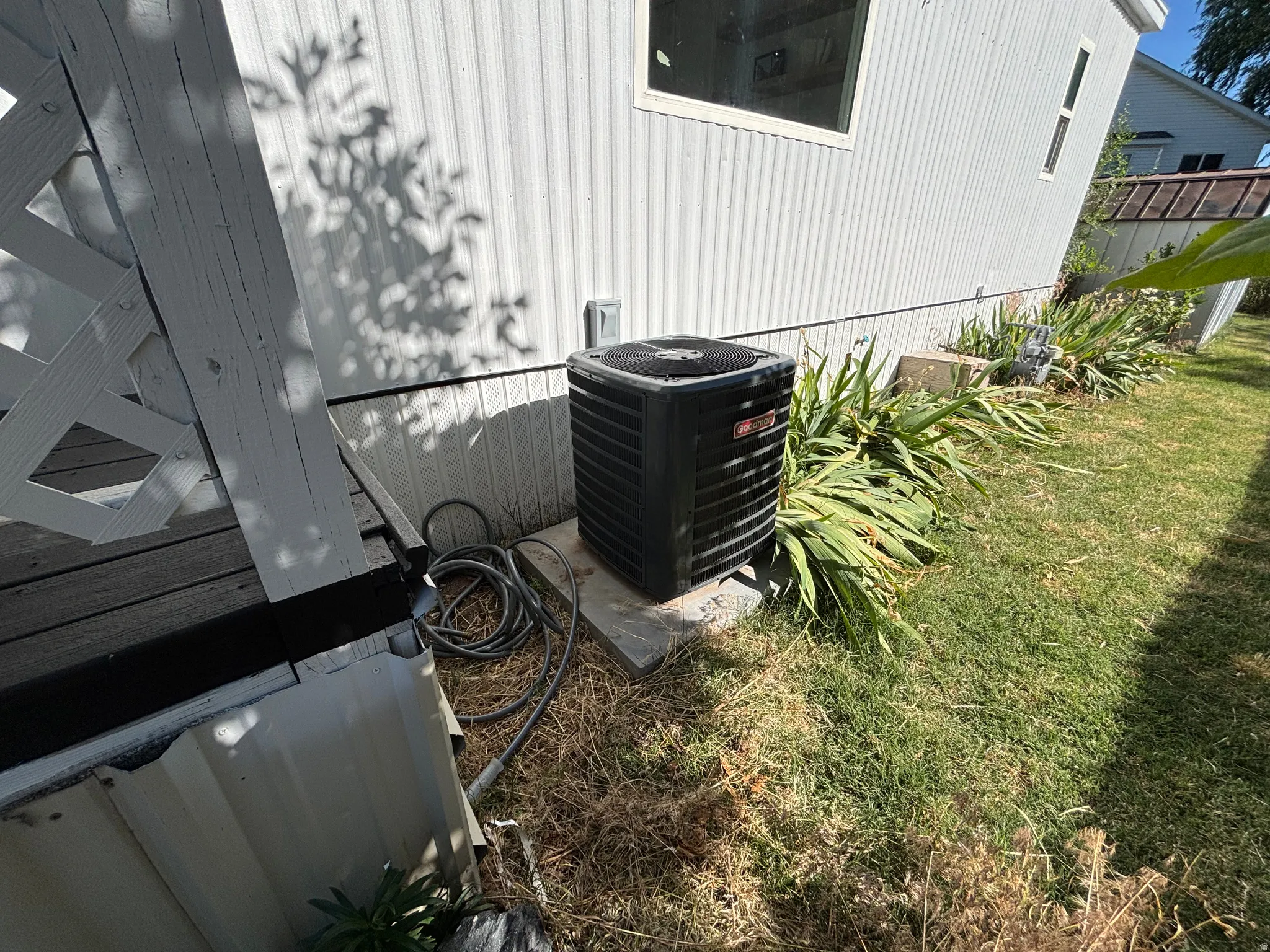 White corrugated siding exterior with a dark-colored outdoor HVAC unit on a concrete pad, surrounded by green landscaping and a wooden deck