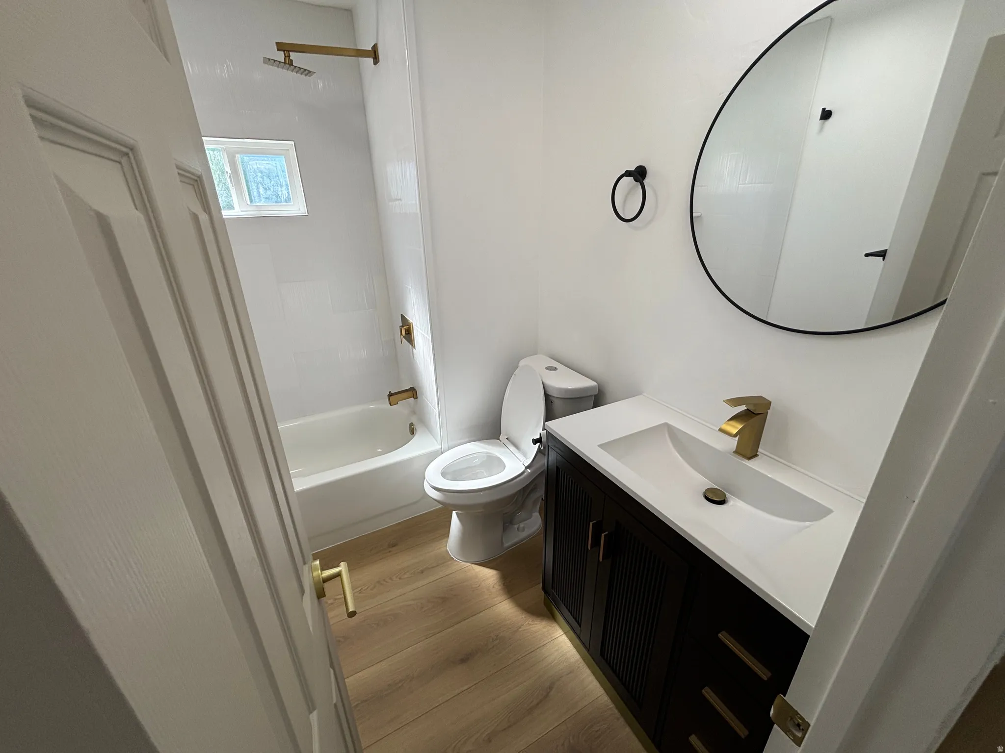 Bathroom featuring a dark wood vanity with a white integrated sink and brass-finish faucet, a circular mirror with a black frame, a white bathtub with subway tile surround and brass-finish fixtures, and wood-finish flooring