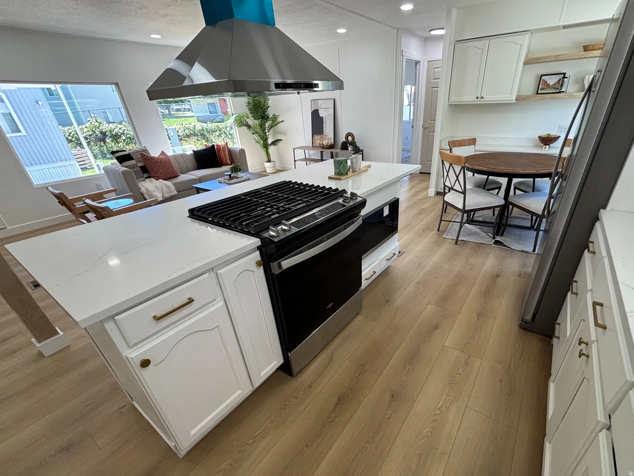 Open-concept kitchen featuring a center island with integrated range, stainless steel vent hood, white cabinetry with brass hardware, and wood-finish flooring