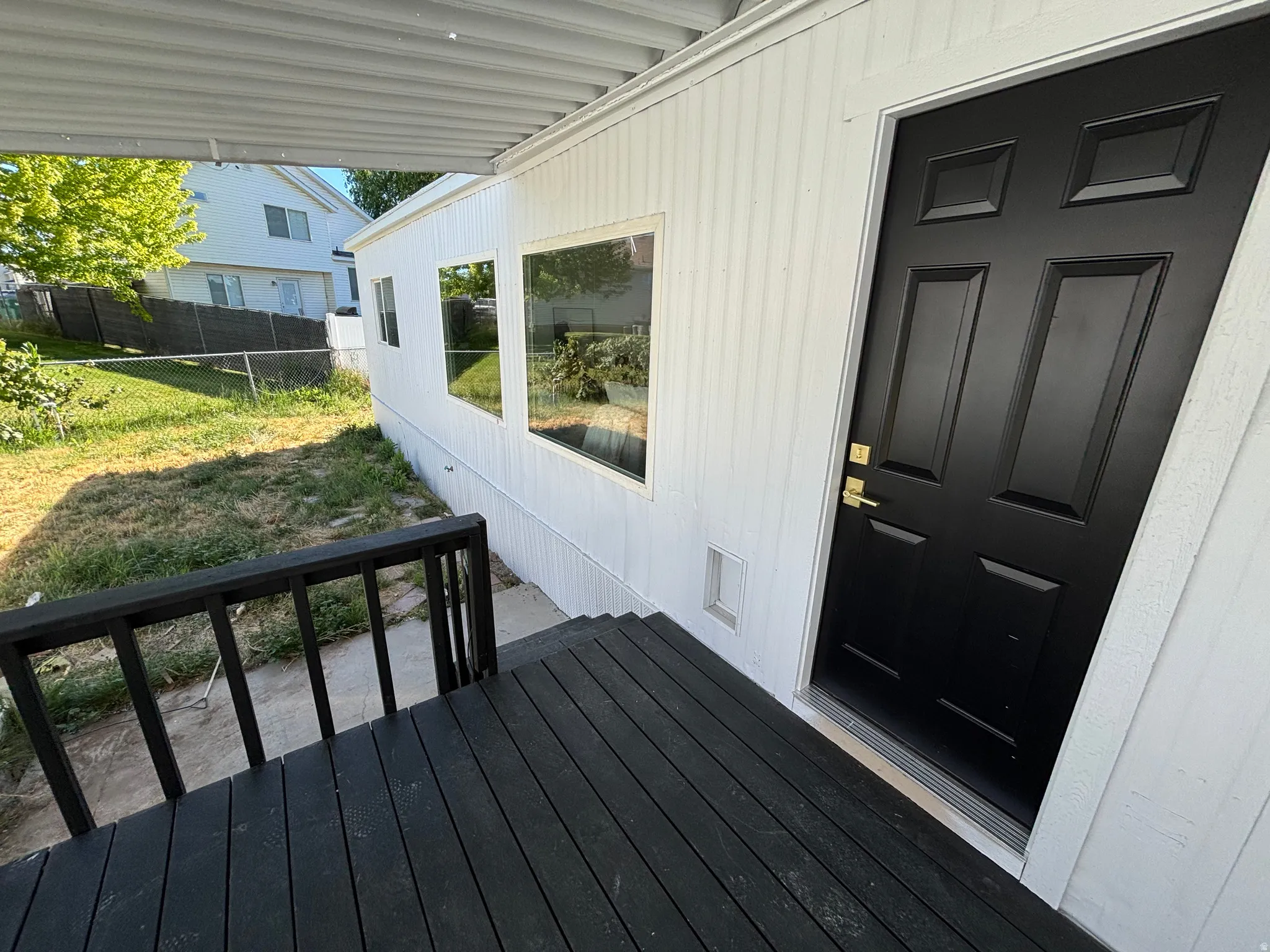 Covered outdoor deck area with dark wood-finish decking and railing, a sleek black entry door with gold-tone hardware, and white vertical siding
