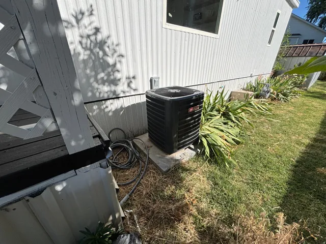 White corrugated siding exterior with a dark-colored outdoor HVAC unit on a concrete pad, surrounded by green landscaping and a wooden deck
