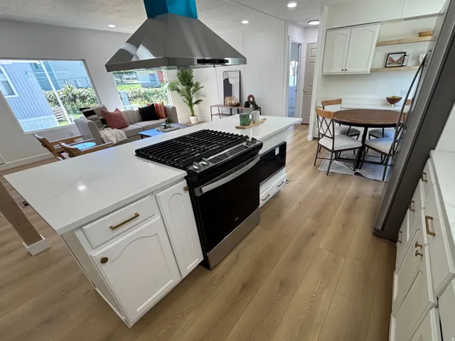 Open-concept kitchen featuring a center island with integrated range, stainless steel vent hood, white cabinetry with brass hardware, and wood-finish flooring