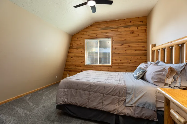 Slanted ceiling room featuring a wood plank accent wall, window with blinds, and a ceiling fan