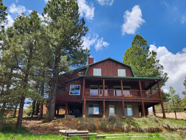 Rustic barn-style home featuring a covered upper-level deck with wood railing, prominent wood siding, a green metal roof, and surrounding mature pine trees
