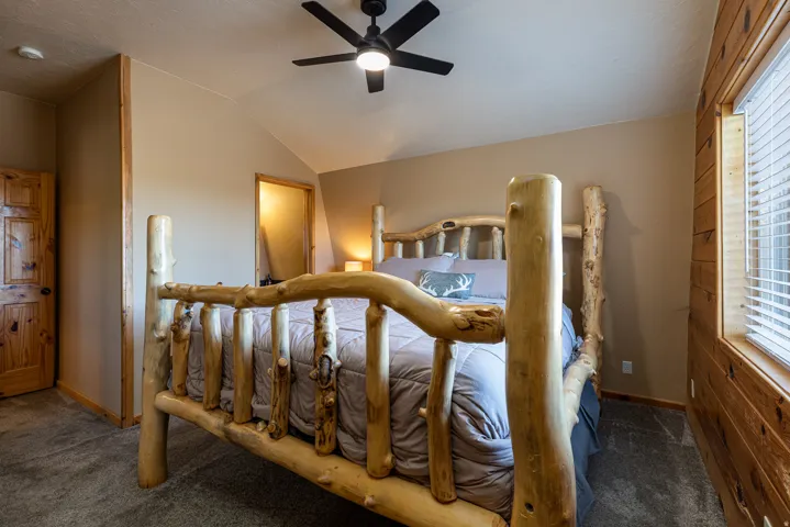 Vaulted ceiling bedroom featuring a ceiling fan, a window with horizontal blinds, wood-paneled accent wall, and a natural wood door has ensuite bath