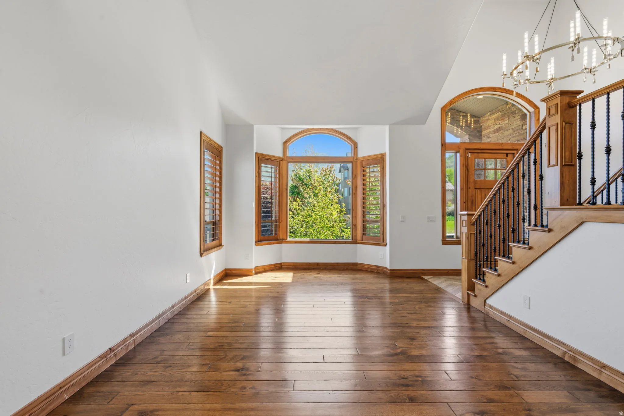 Hardwood flooring throughout the entry, featuring a curved bay window with plantation shutters, a grand staircase with wrought iron balusters, a wood-finish front door with an arched transom, and an ornate chandelier