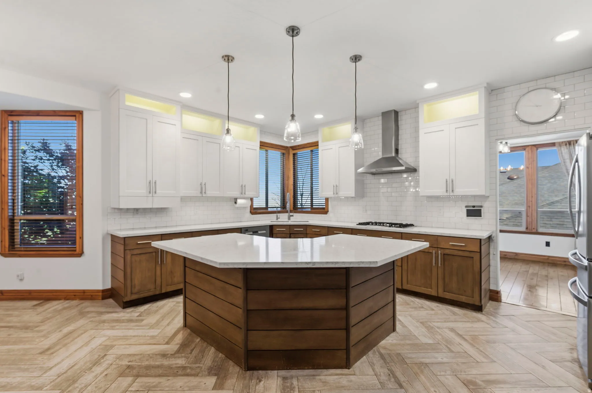 Kitchen featuring a multi-sided island with a light-toned countertop, two-tone cabinetry, white subway tile backsplash, stainless steel range hood, and herringbone pattern tile flooring.
