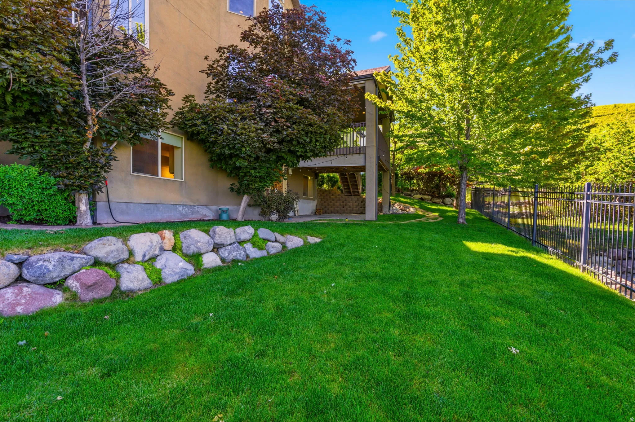Expansive green lawn featuring mature trees, a stone retaining wall, and a black metal fence