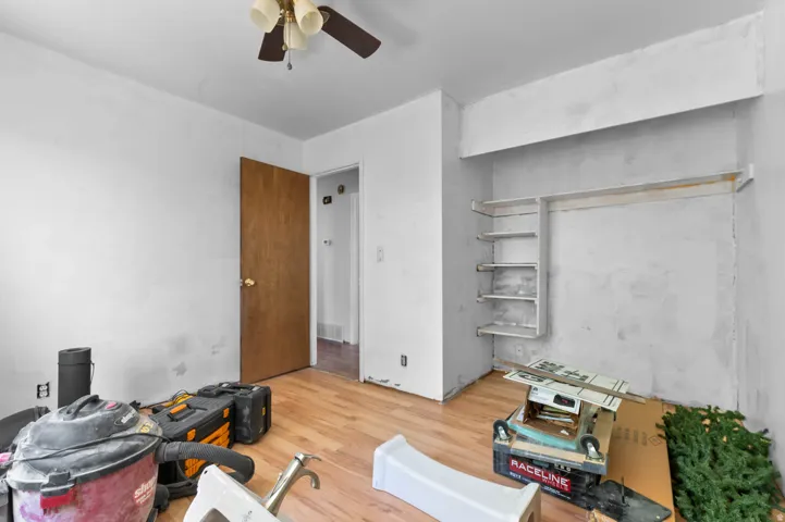 Room featuring wood-finish flooring, a ceiling fan with integrated lighting, a wooden interior door, and an integrated wall alcove with shelving