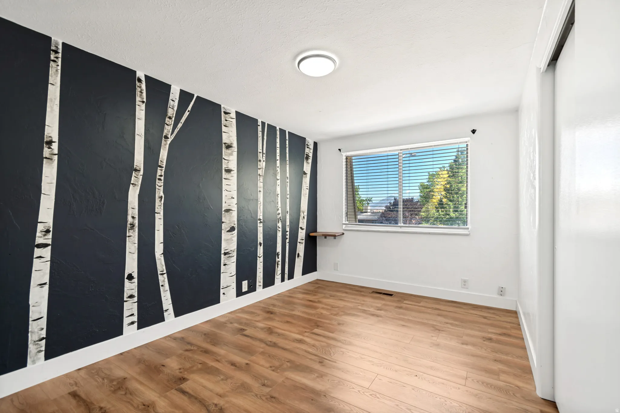 Room featuring a dark accent wall with birch tree motif, wood-finish flooring, a window with blinds, a white ceiling, and a flush-mount ceiling light fixture