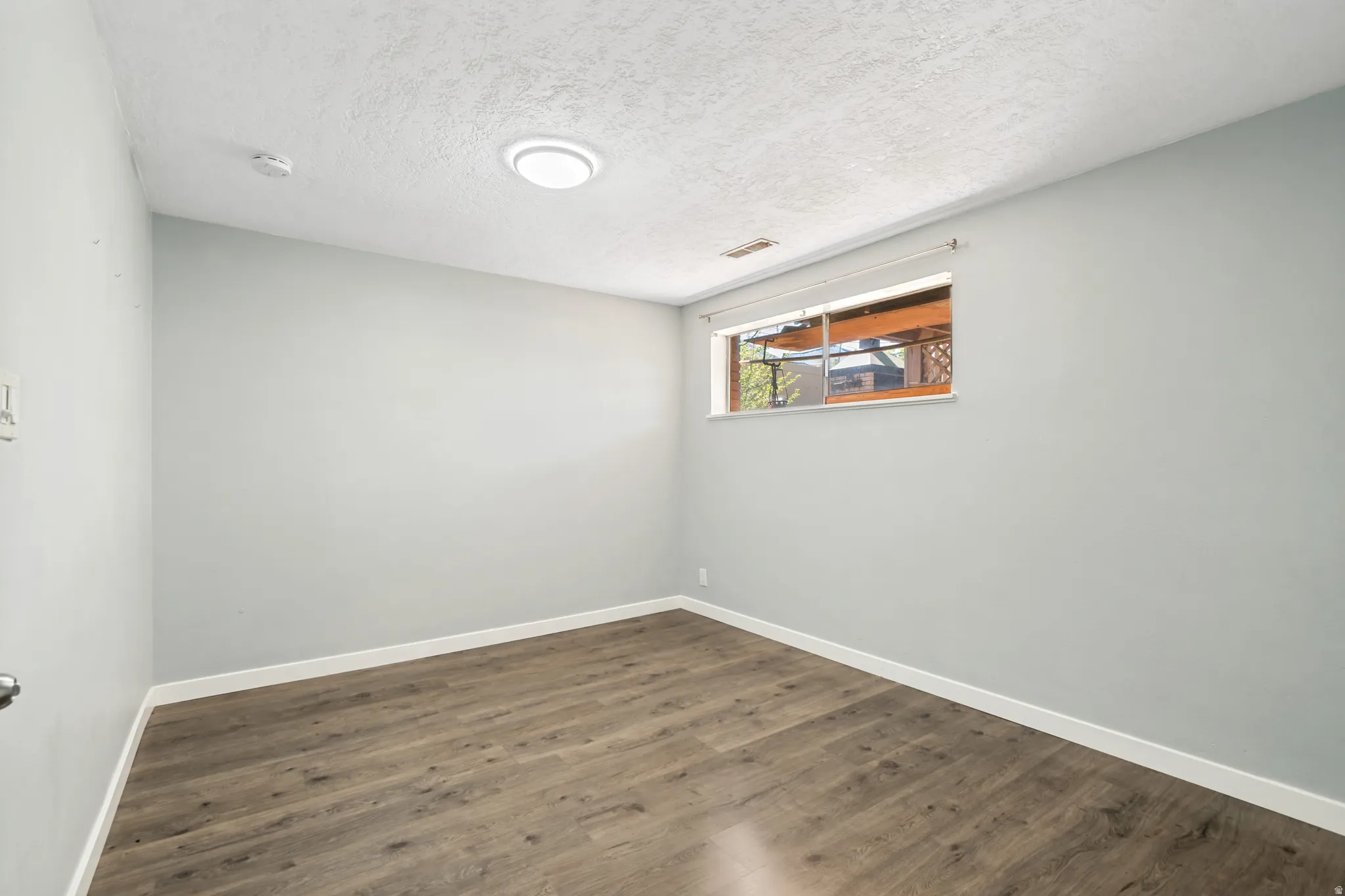Spacious room featuring wood-finish flooring, light grey painted walls, white baseboards, and a window with natural light