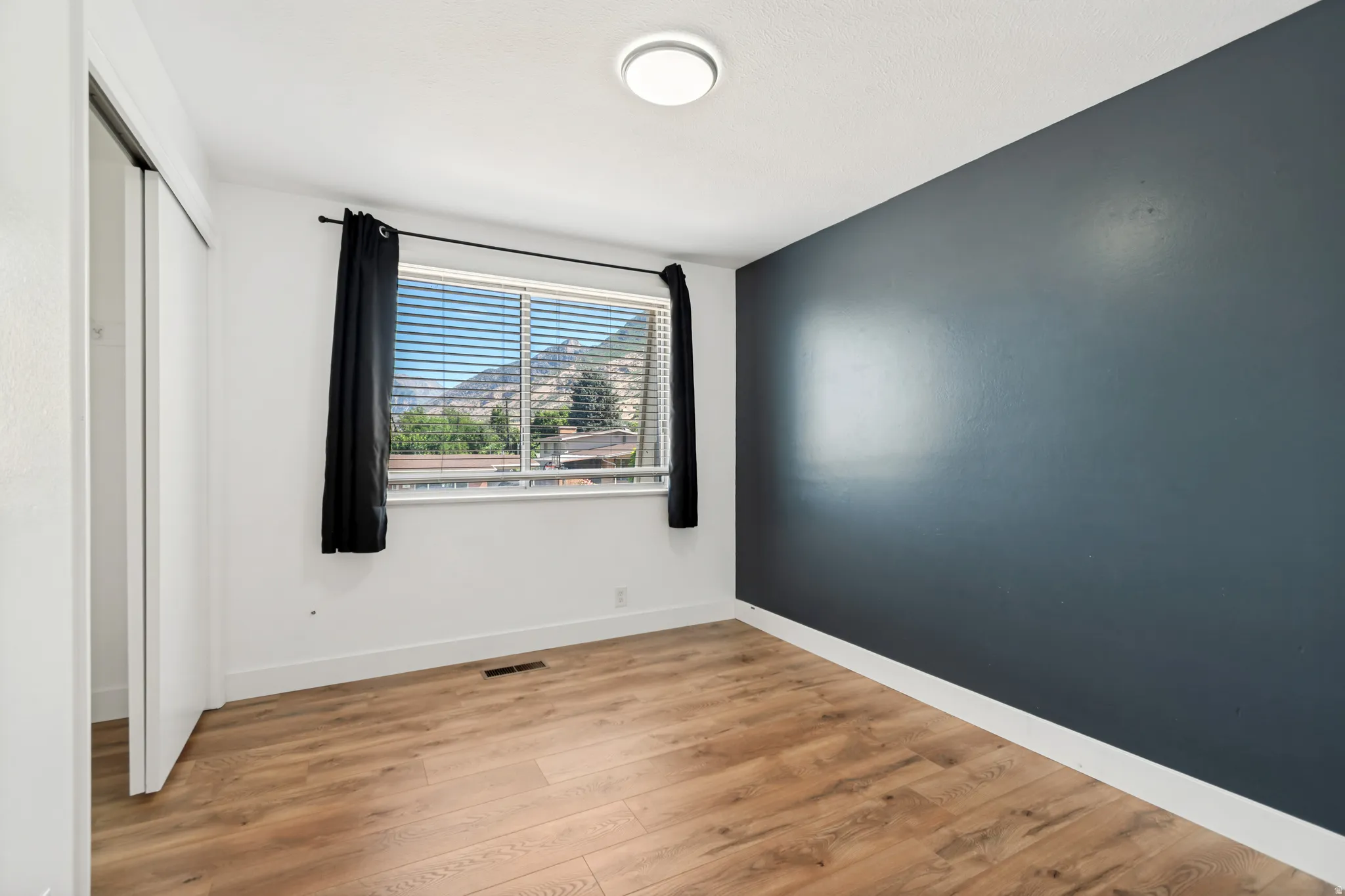 Room featuring a window with integrated blinds, wood-finish flooring, a contemporary flush-mount ceiling light, a built-in closet with sliding doors, and a contrasting accent wall