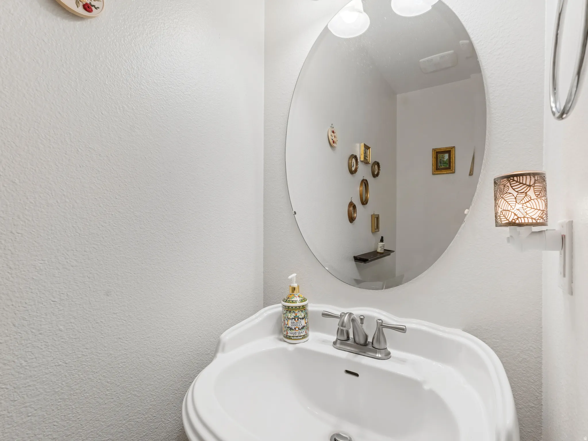 Pedestal sink with a dual-handle faucet, oval mirror, wall-mounted light fixture, and white textured walls