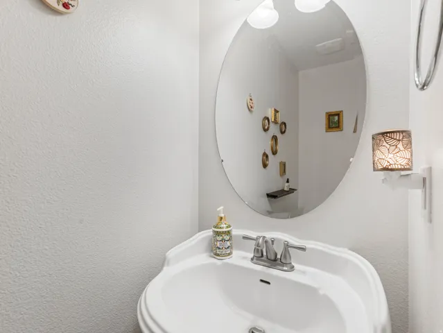 Pedestal sink with a dual-handle faucet, oval mirror, wall-mounted light fixture, and white textured walls