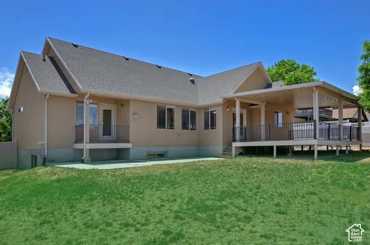 Expansive rear exterior featuring a covered deck, stucco siding, multiple windows, a ground-level patio, and a manicured lawn