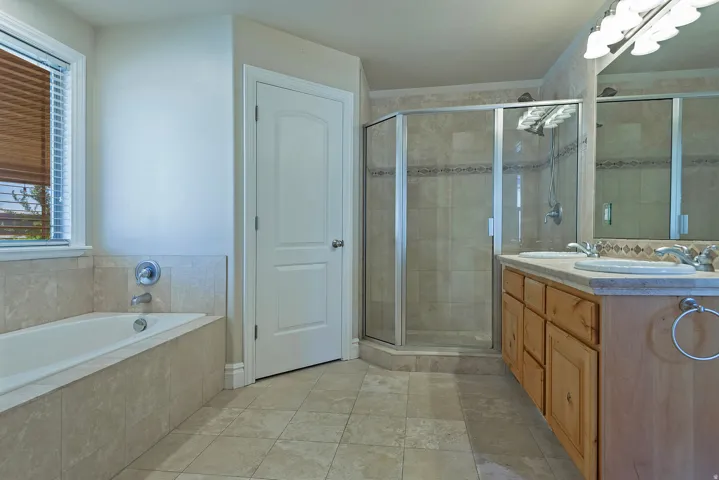 Bathroom featuring a built-in tub with surrounding tile, a separate glass-enclosed shower, a double vanity with undermount sinks and light wood cabinetry, and a window with blinds