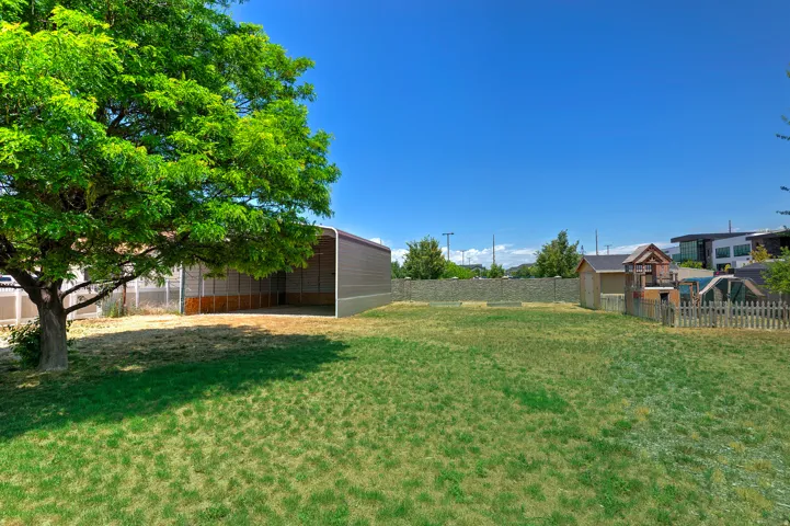 Expansive backyard featuring a large metal carport structure, mature shade tree, and a wooden picket fence