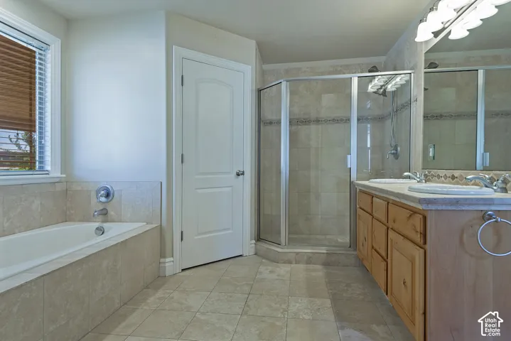 Bathroom featuring a built-in tub with surrounding tile, a separate glass-enclosed shower, a double vanity with undermount sinks and light wood cabinetry, and a window with blinds