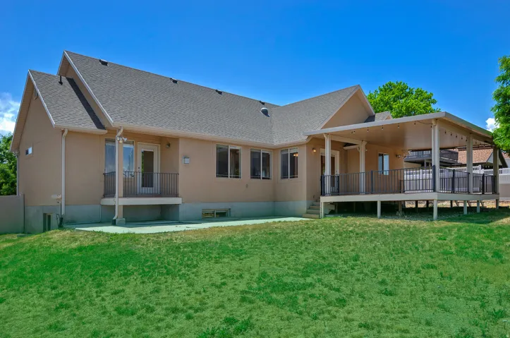 Expansive rear exterior featuring a covered deck, stucco siding, multiple windows, a ground-level patio, and a manicured lawn