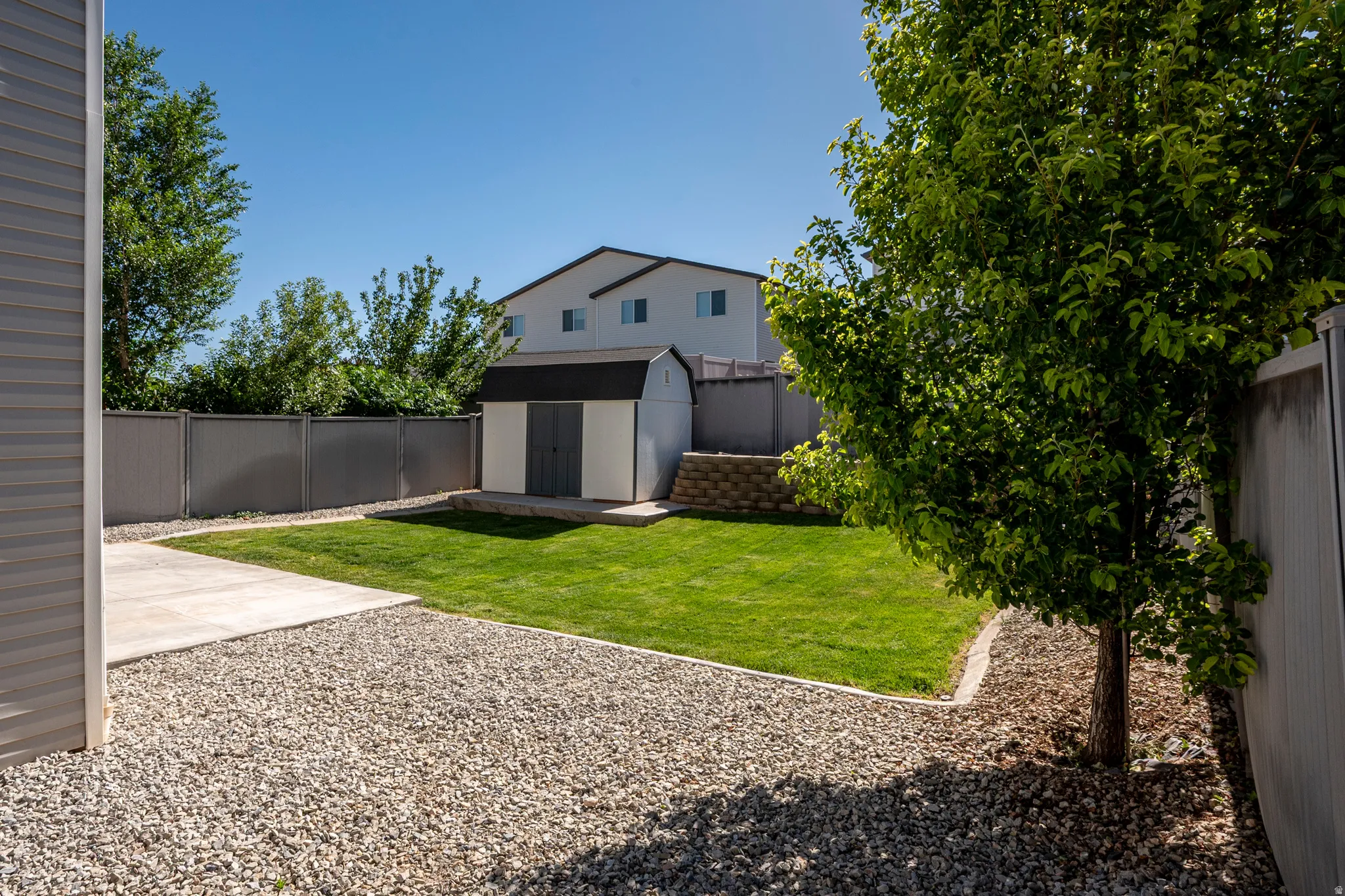 Expansive backyard featuring a lush lawn, a detached storage shed, a gravel ground cover, a concrete patio, and a retaining wall
