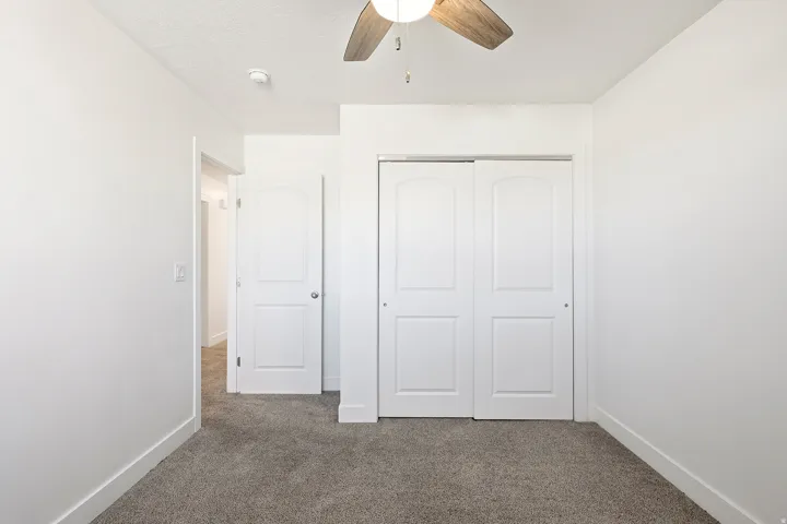 Interior room featuring neutral wall paint, gray carpeting, a ceiling fan with wood-finish blades, white paneled doors, and white baseboards