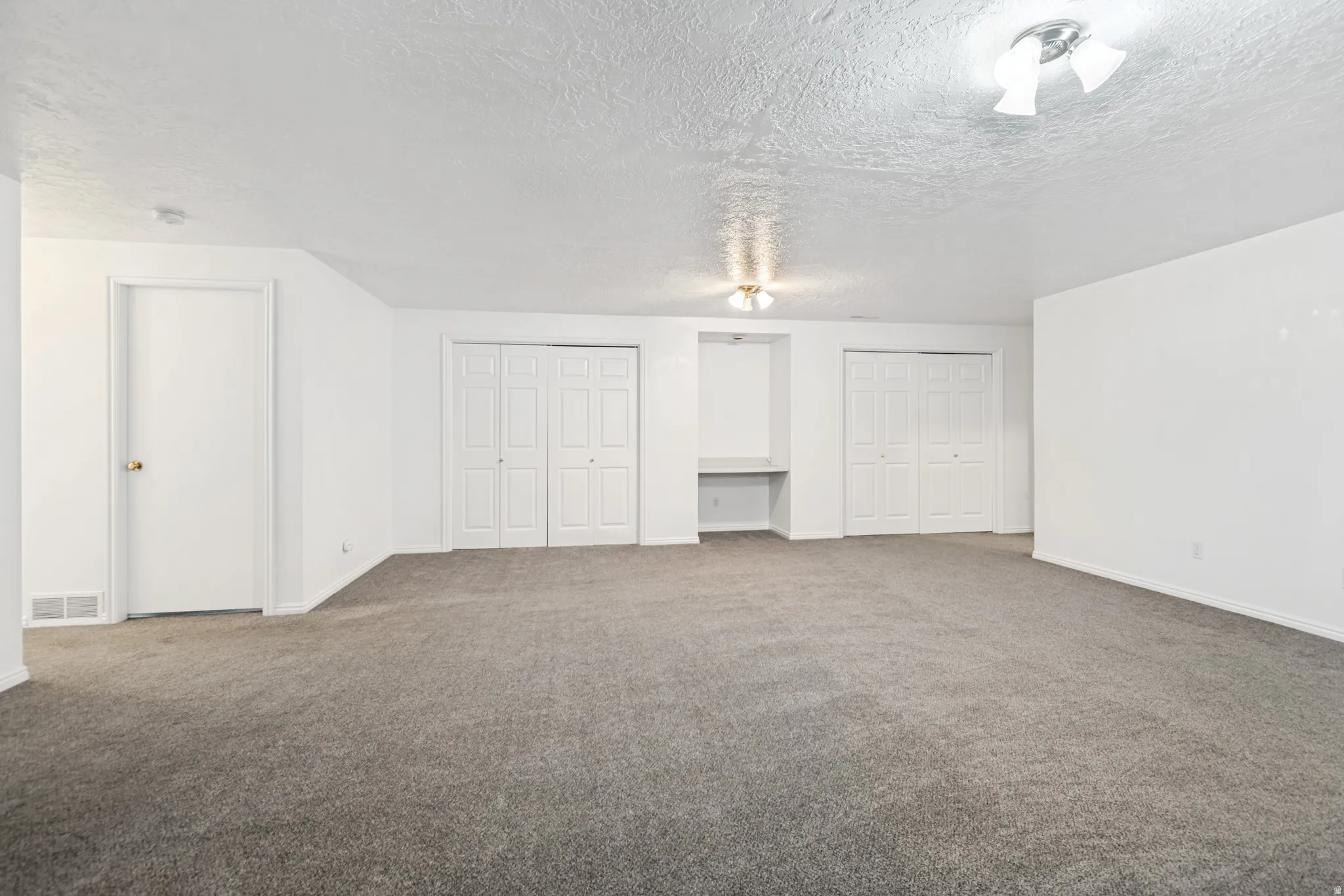 Spacious carpeted room featuring two sets of white bi-fold closet doors, a built-in desk niche, and overhead ceiling lighting