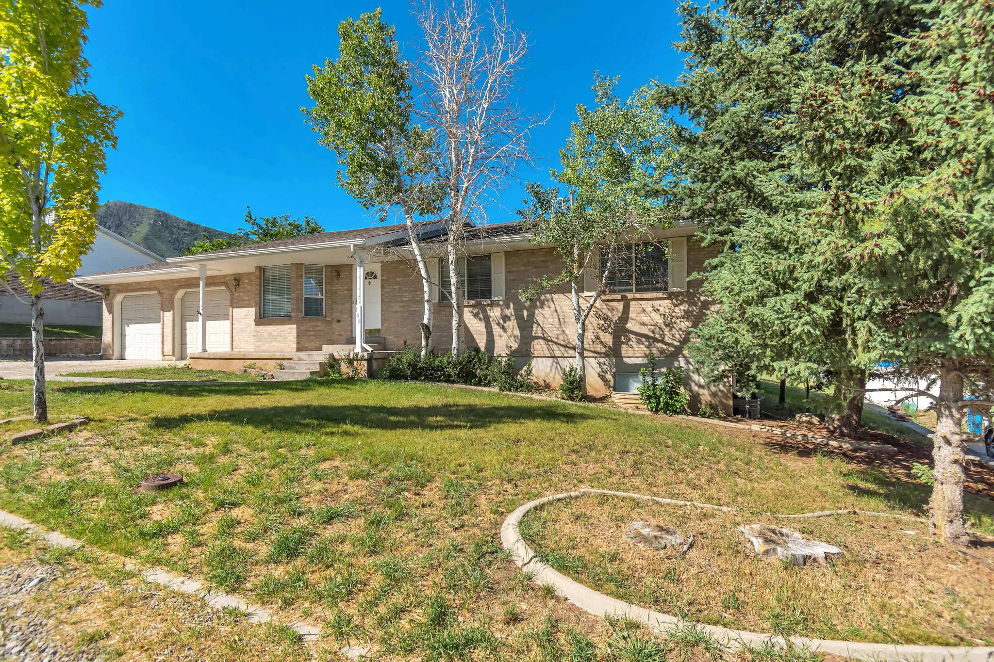 Brick exterior property with an attached two-car garage, mature trees, a gently sloped front lawn, a concrete border, and a mountain backdrop
