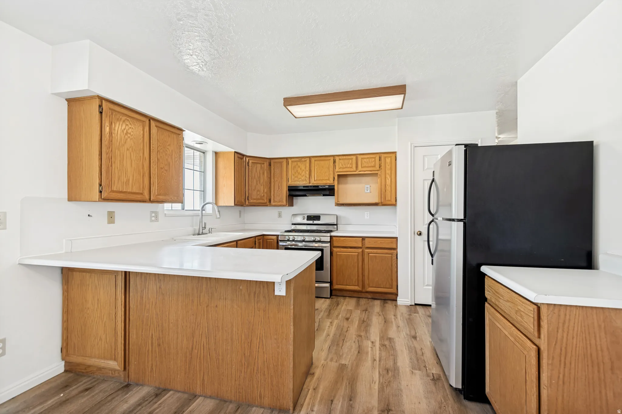 Open-concept kitchen featuring light wood cabinetry, white countertops, stainless steel appliances, wood-finish flooring, and a large window above the sink