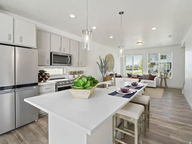 Open-concept kitchen featuring light-toned cabinetry, stainless steel appliances, a kitchen island with seating, pendant lighting, and wood-finish flooring